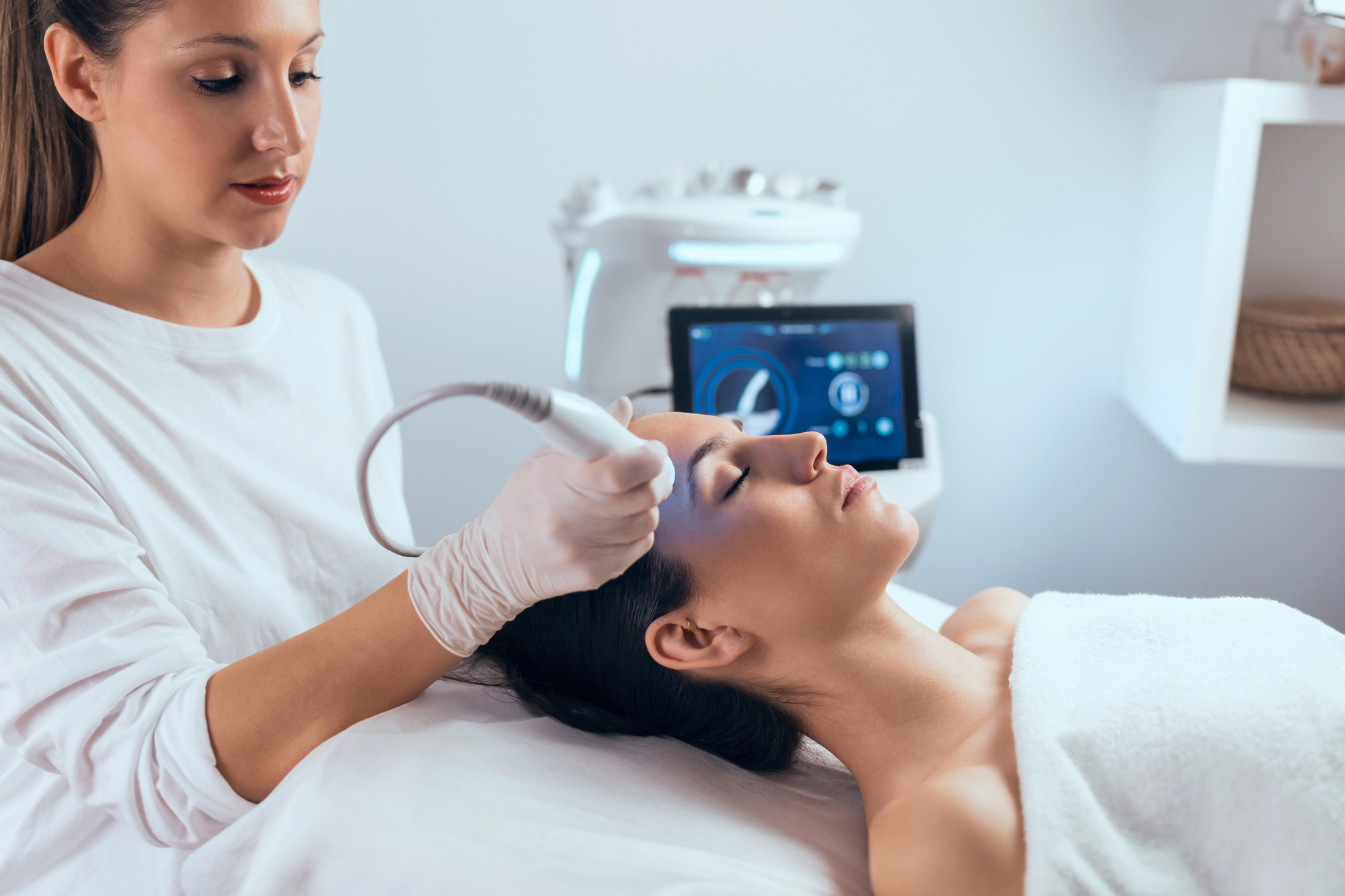 Aesthetician performs a laser spa treatment to a woman lying on clinic bed, with machine in the background